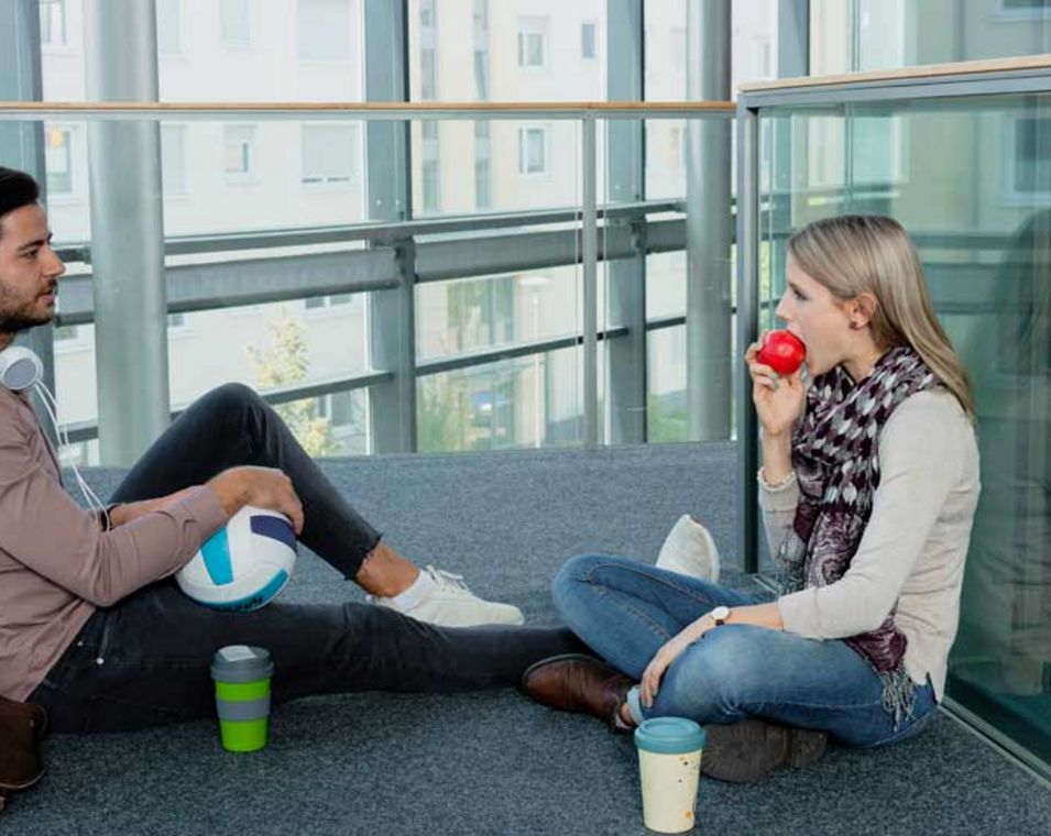 Junge Frau und junger Mann in modernem Bürogebäude machen Pause mit Kaffee, Apfel, Volleyball und Musik