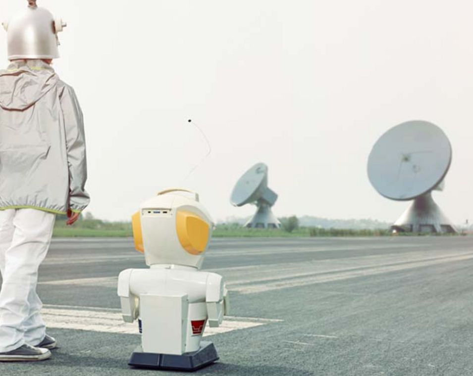 Als Roboter verkleideter Mensch und ein Roboter stehen auf einem Rollfeld mit Satellitenschüsseln