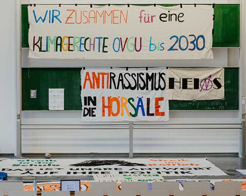 AUfnahme aus dem Hörsaal der Universität Magdeburg, an der Tafel hängen Banner "Wir zusammen für eine klimagerechte OVGU bis 2030" und "Antirassismus in die Hörsäle".