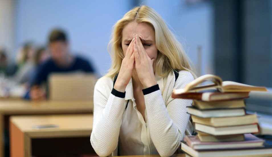 Eine Studentin sitzt an einem Bibliothektsarbeitsplatz und ist verzweifelt, sie hebt die Hände vor ihr Gesicht.