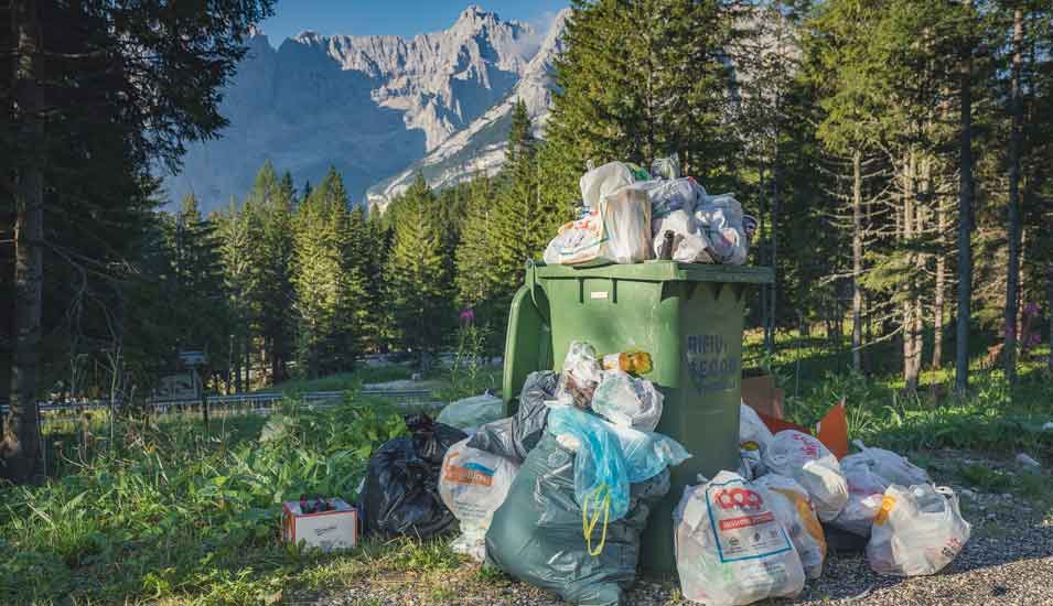 Überlaufende Mülltonne und Müllsäcke an einem Weg in den Dolomiten, die man im Bildhintergrund erkennen kann.